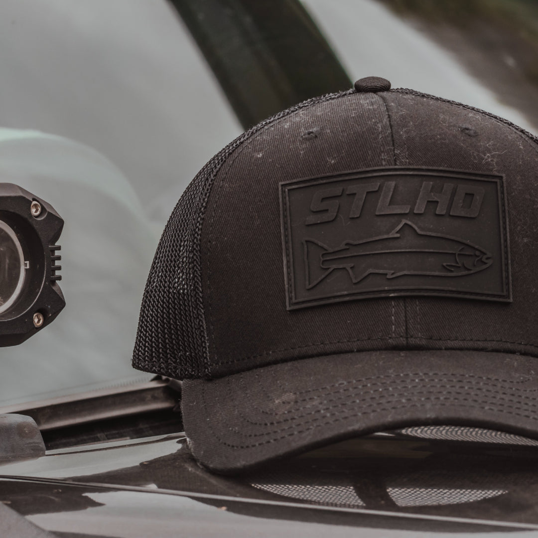 All-black STLHD Blackout Trucker hat with black logo on the front panel and black mesh back.