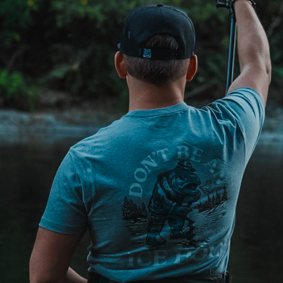 A man viewed from behind wearing a teal blue t-shirt with a large back graphic of a fisherman and the text 'DON'T BE A IDLE PLOUGH'.