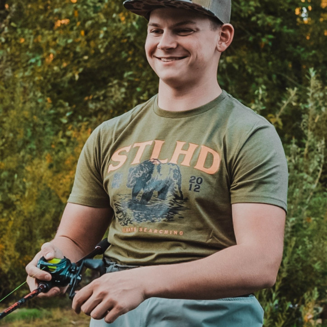A smiling man outdoors wearing an olive green graphic t-shirt with a bison print and the text 'STLHDO' and 'TIME SEARCHING', holding a fishing rod. He is wearing a camouflage cap and light blue pants, with a blurred background of green foliage.