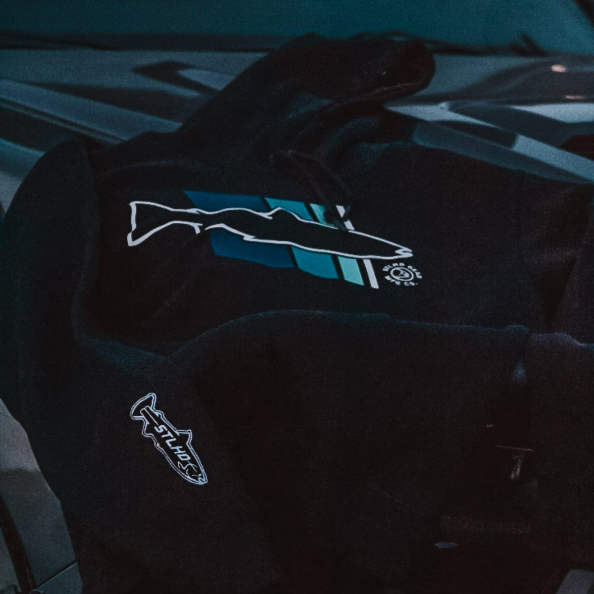 A black hoodie featuring a stylized fish graphic on the chest in white and teal-blue, with a smaller matching fish logo patch on the sleeve, photographed in a dark moody setting.