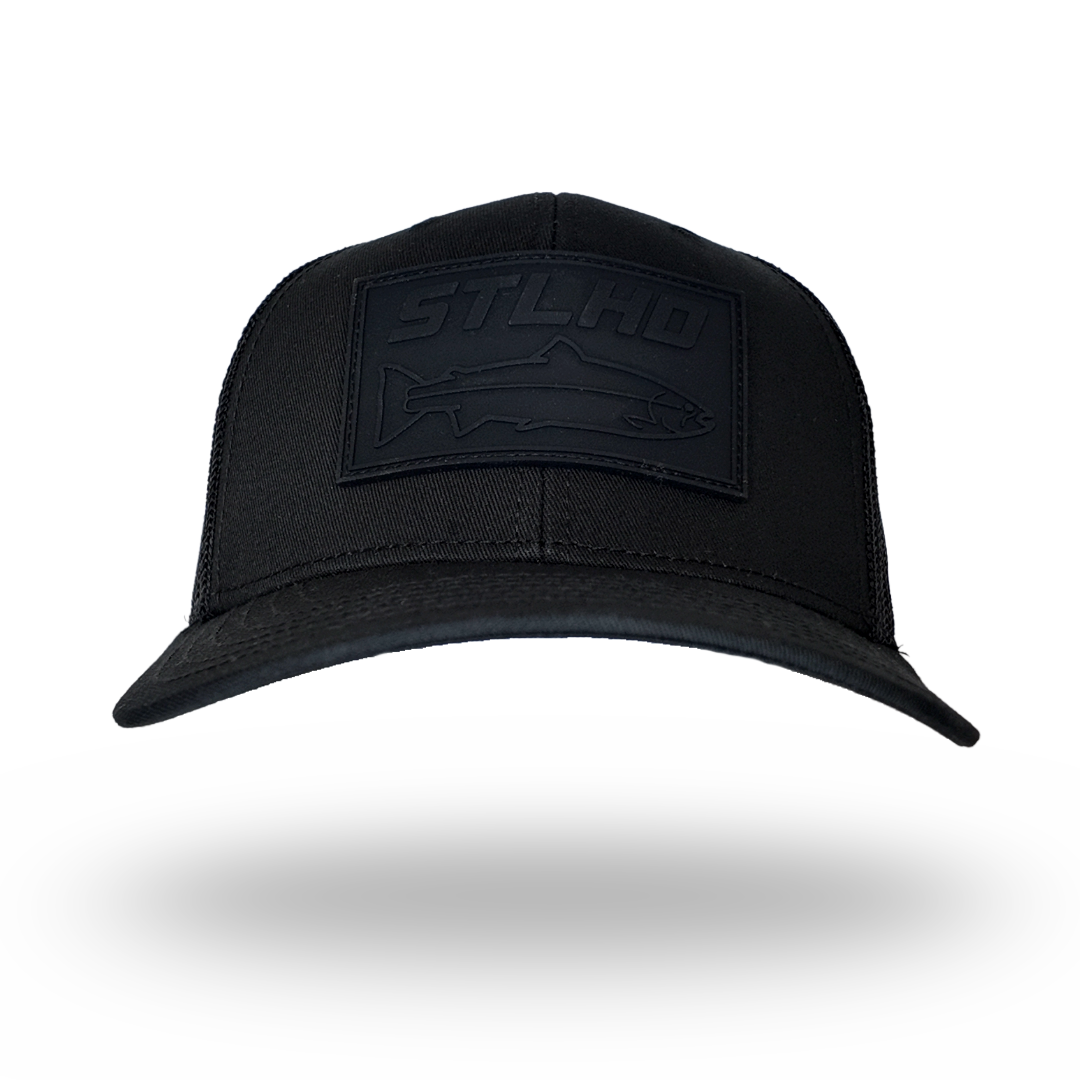 A black trucker-style cap with a tonal black rectangular patch on the front featuring the embossed brand name 'STLHO' and a fish silhouette, shown against a plain white background with a soft shadow underneath.