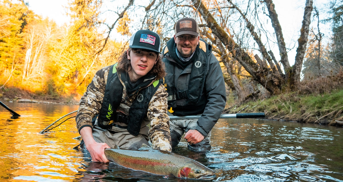 Two people kneeling in a river holding a large rainbow trout, with autumn trees in the background. The person in front wears a black cap with a US flag, a camouflage jacket, and a black life vest, while the person behind wears a brown cap and a dark gray jacket.