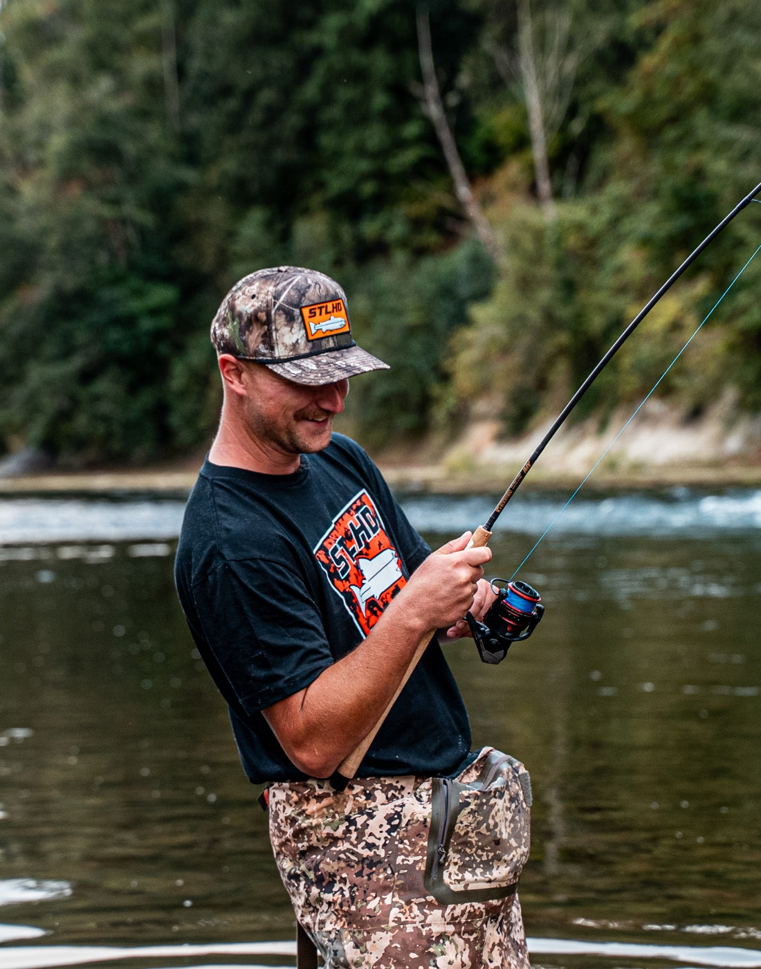 A man wearing a camouflage cap, black graphic t-shirt, and camouflage pants stands in a river holding a fishing rod, with a forested riverbank in the background.