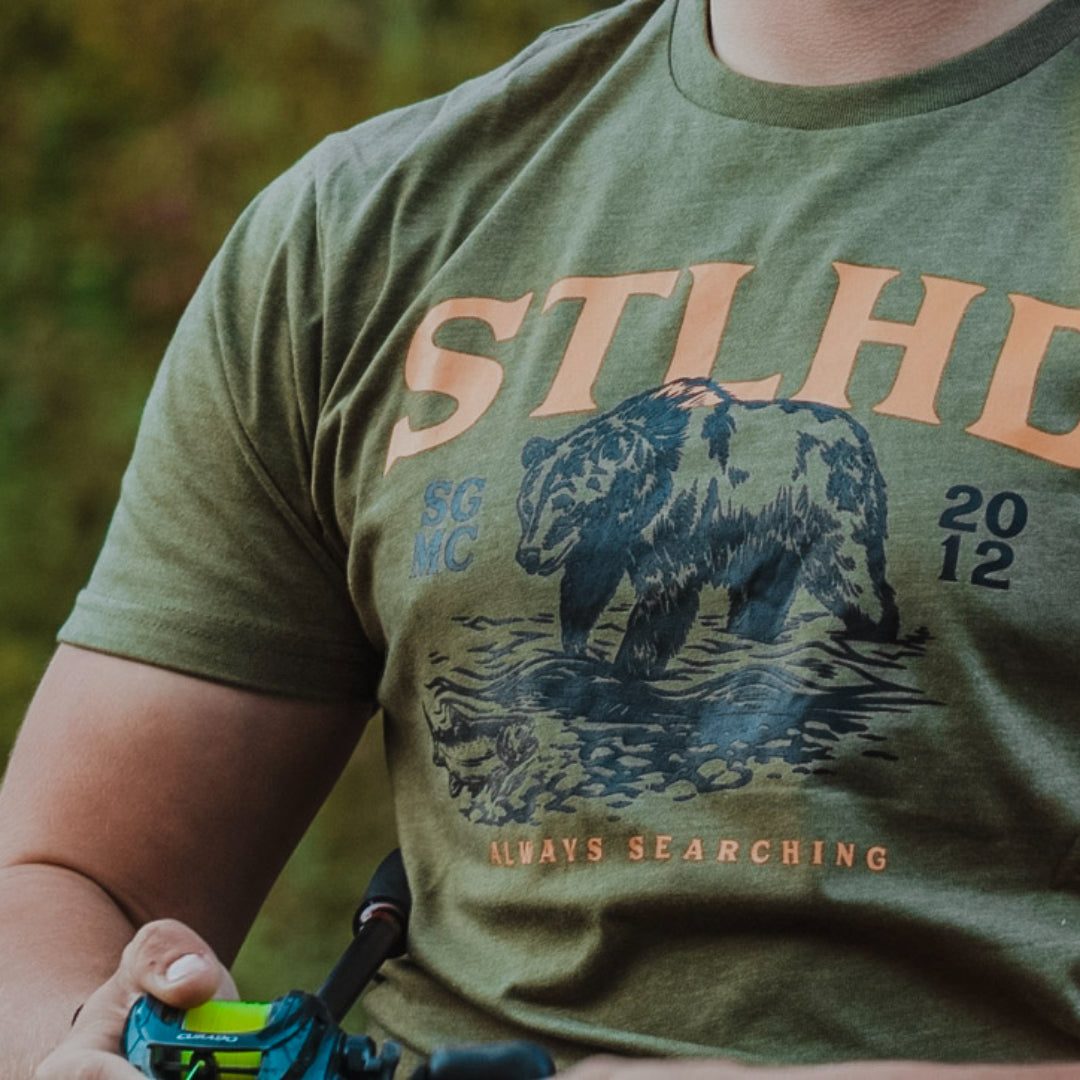 Close-up of a person wearing an olive green t-shirt with a graphic print of a grizzly bear standing in water, with the text 'STLH' and 'ALWAYS SEARCHING' in orange, and 'SC NC 2012' in blue.