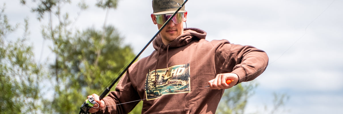 A man wearing a brown hoodie with a large graphic print on the front, sunglasses, and a cap, holding a fishing rod outdoors with trees and sky in the background.