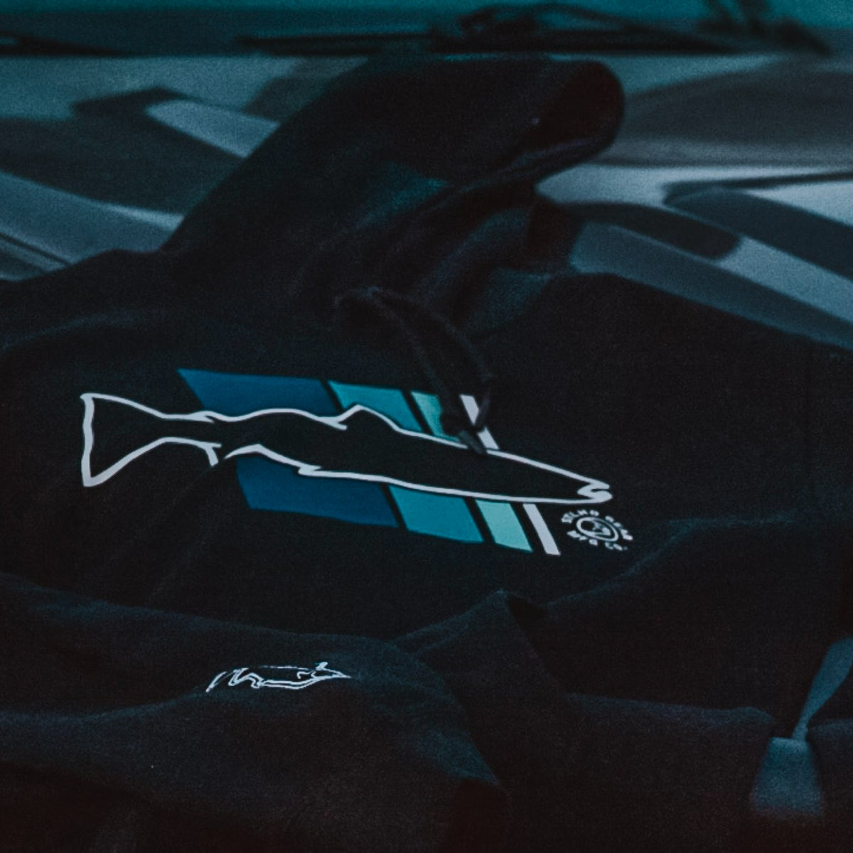 A dark navy or black hoodie laid flat, featuring a stylized fish graphic with blue and teal stripes outlined in white on the chest.