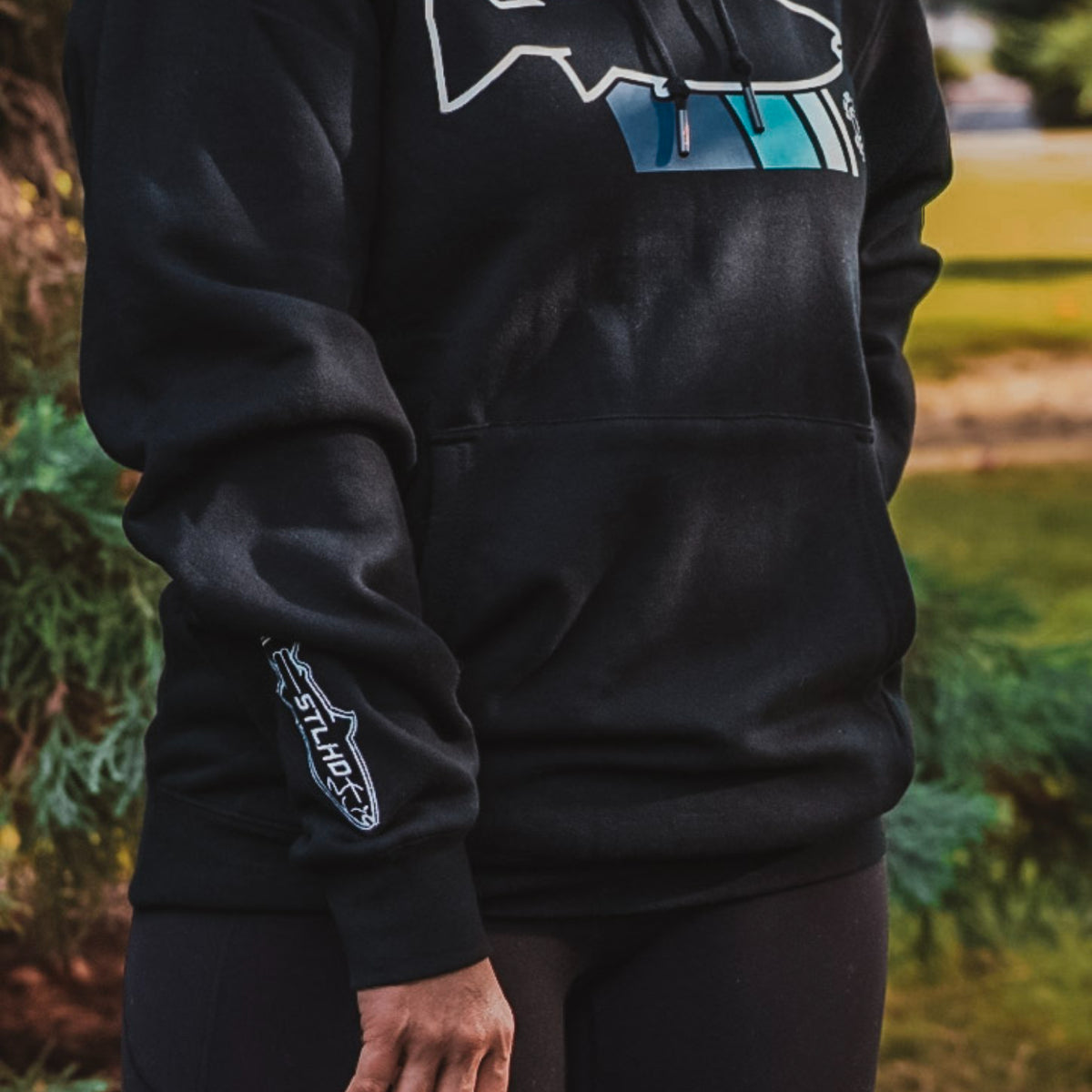 A person wearing a black pullover hoodie with a white outlined fish graphic and blue accents on the chest, plus a white 'STLHD' logo on the left sleeve, photographed outdoors with blurred greenery in the background.