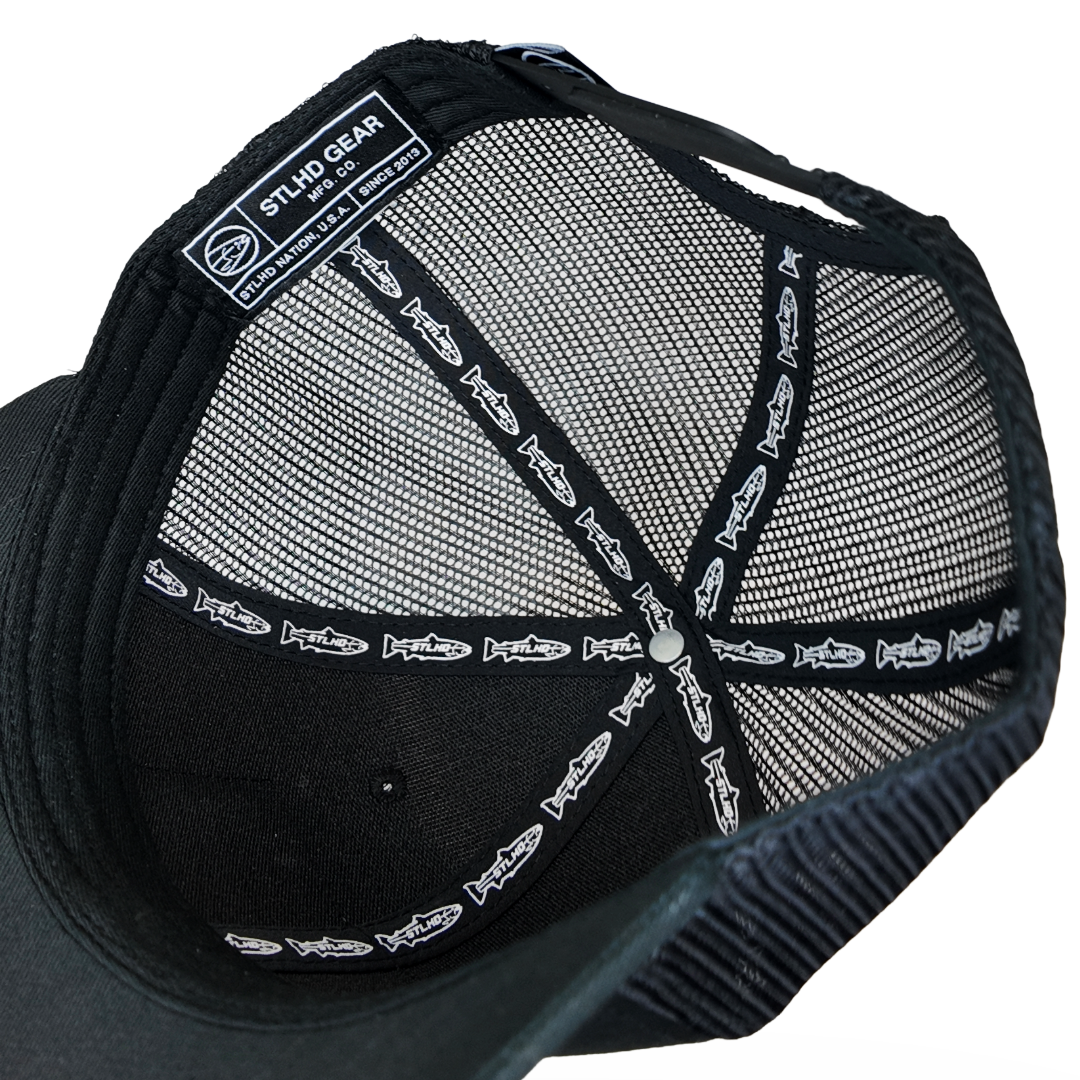 Close-up of the inside of a black trucker-style cap, showing a black mesh back, black fabric crown, printed fish logo trim on the suspension straps, and a black interior patch that reads 'STUD.GEAR' with smaller text below.